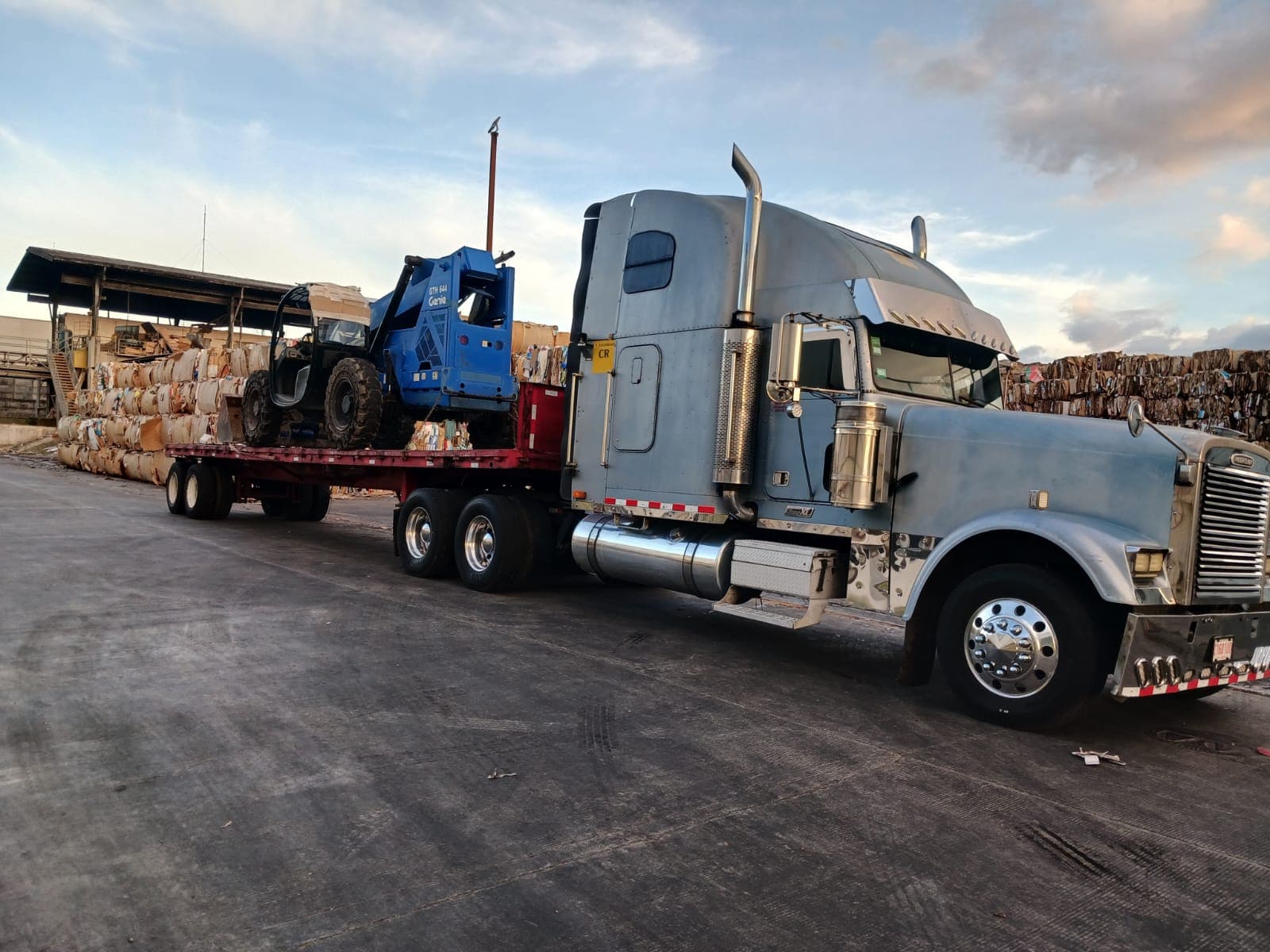 Freightliner plateado con un telehandler Genie en la plataforma, al atardecer