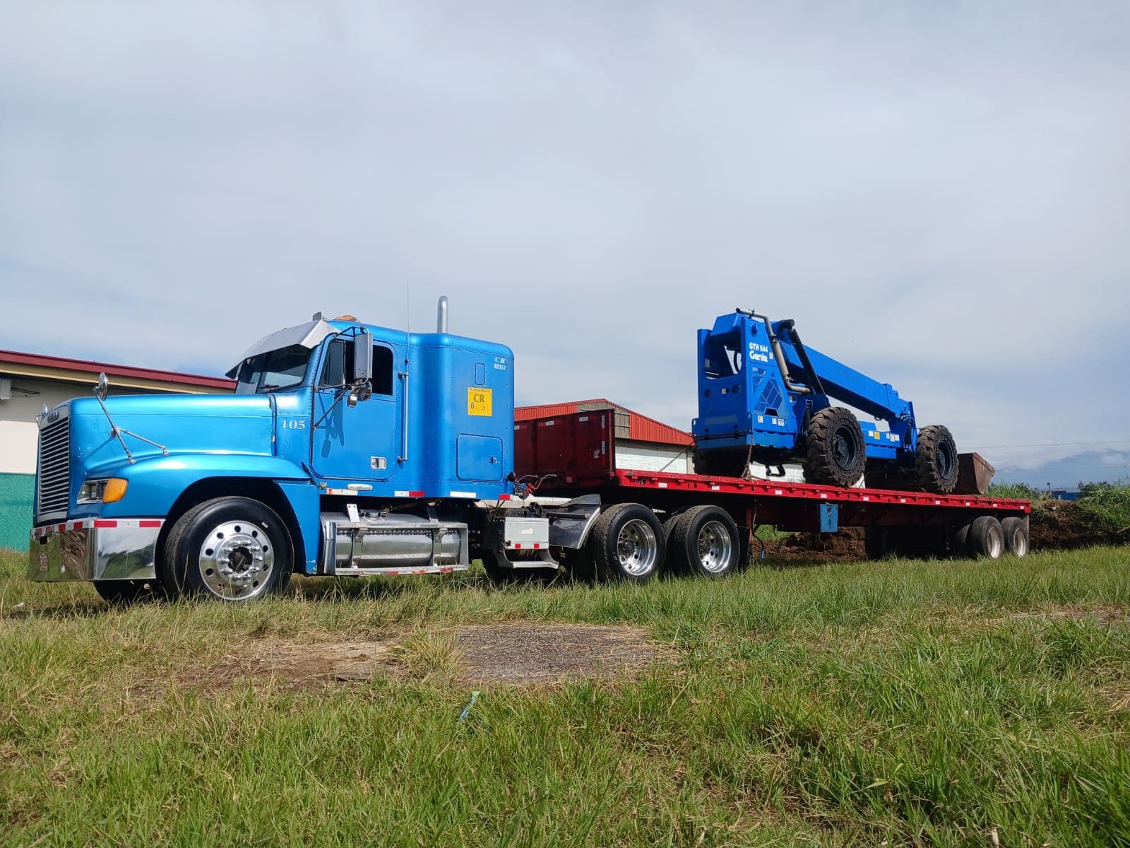 Cabezal Freightliner azul con un telehandler Genie amarrado en la plataforma